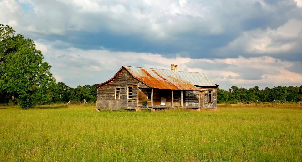 A weathered farmhouse stands in a vibrant rural field under a cloudy sky.